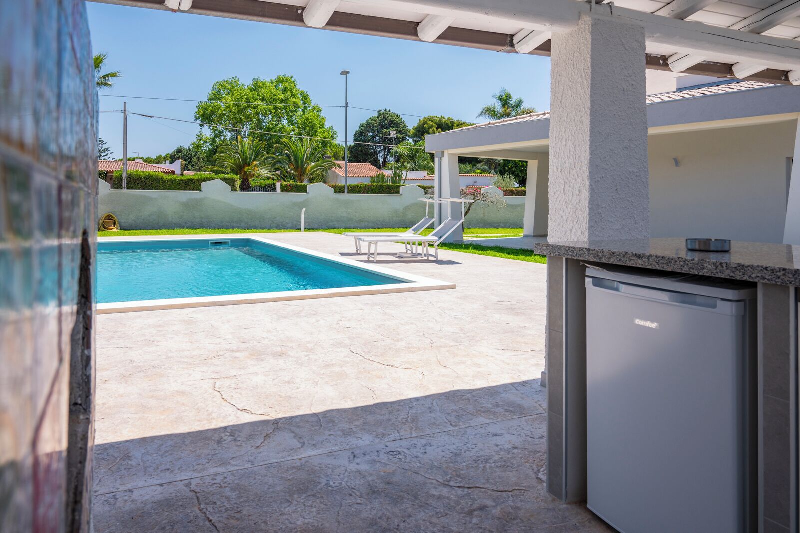 Zona de piscina con suelo de hormigón, piscina y área de estar cubierta con sillas.