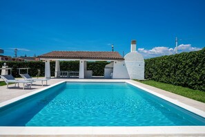 Et rektangulært swimmingpool med klart, blåt vand, omgivet af en hvid poolterrasse. Der er liggestole og et overdækket siddeområde med et flisebelagt tag. En hvid stensøjle er synlig til højre for poolen.