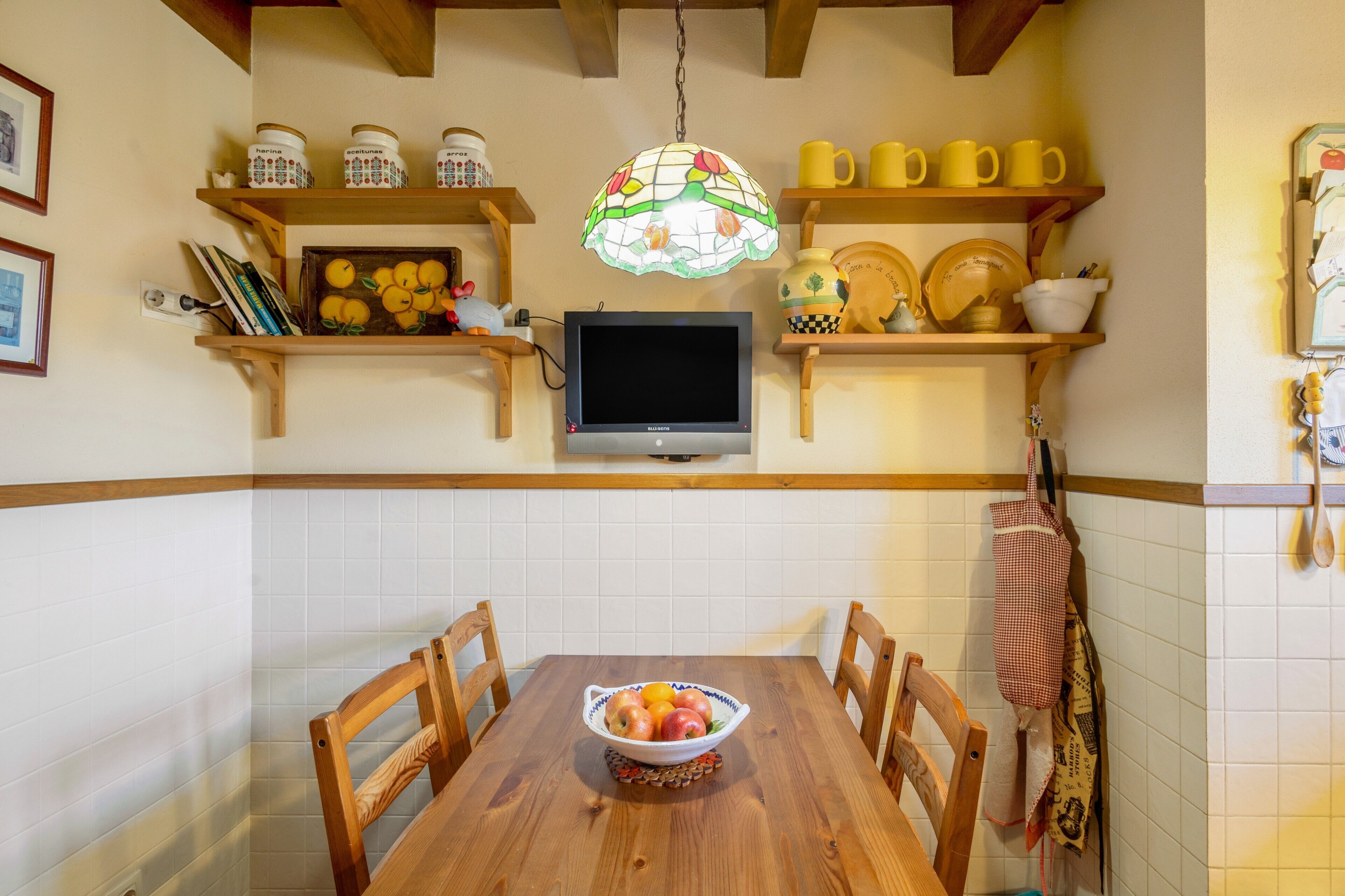 Una cocina con una mesa de madera, sillas, un tazón de fruta, un televisor y estantes con utensilios de cocina.