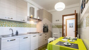 A compact kitchen with a sink, stove, and microwave. There is a dining table with a green tablecloth and white chairs. - Homiday - Jasmine One Min[Hospital][Airport-Free Parking] (Pisa)