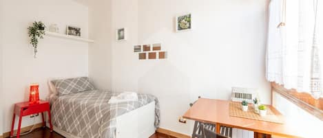 A bedroom with a bed, a red nightstand, a wooden table with chairs, and a window with curtains.
