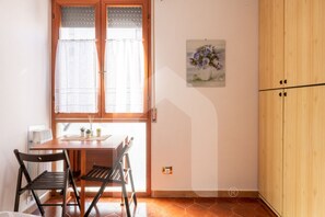A dining area with a wooden table and chairs, a window with curtains, a wall-mounted picture, and a wooden wardrobe. - Homiday - Jasmine, 1 min away [Hospital][Airport—Free Parking] (Pisa)