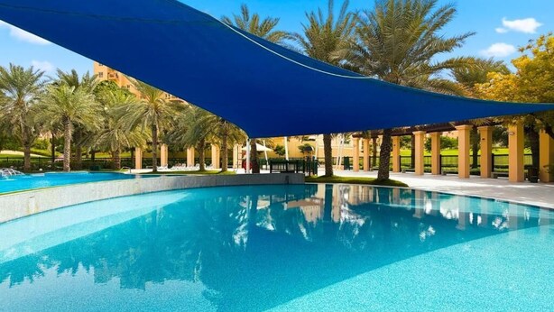 Una piscina azul cristalina con un toldo azul, rodeada de palmeras y un edificio al fondo.