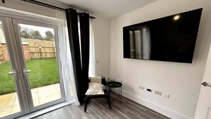 Ein Zimmer mit einer Schiebeglastür, die auf eine Terrasse führt, einem schwarzen Sessel, einem kleinen Tisch und einem an der Wand montierten Fernseher.