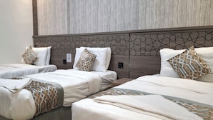 Une chambre d’hôtel avec deux lits simples et un lit plus grand, tous dotés de draps blancs et d’oreillers à motifs.