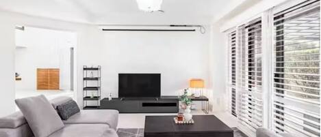 A modern living room with a grey sofa, a black coffee table, a flat-screen TV, and a window with blinds.
