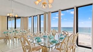 Un comedor con mesa y sillas de vidrio, ventanas amplias con vista a la playa y una lámpara de techo.