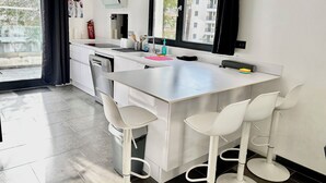 Een moderne keuken met een centraal eiland, witte barstoelen, een vaatwasser en een raam met gordijnen.