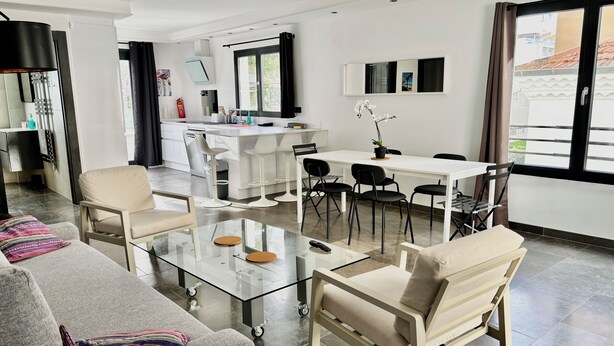 Een moderne woonkamer met een glazen salontafel, een bank met kleurrijke kussens, en een eetgedeelte met een witte tafel en zwarte stoelen.
