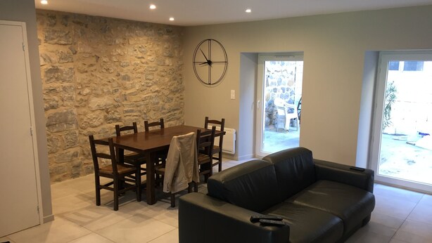 A living room with a stone wall, a dining table with chairs, a black leather sofa, and a large window. - Parenthèse nature au cœur des Gorges du Tarn - Charming house & patio (Rivière-sur-Tarn)