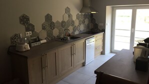 A kitchen with a modern design, featuring a large island with a built-in dishwasher, a gas range, and a stainless steel hood. The backsplash has a geometric tile pattern, and there is a sliding glass door leading to a balcony. - Parenthèse nature au cœur des Gorges du Tarn - Charming house & patio (Rivière-sur-Tarn)