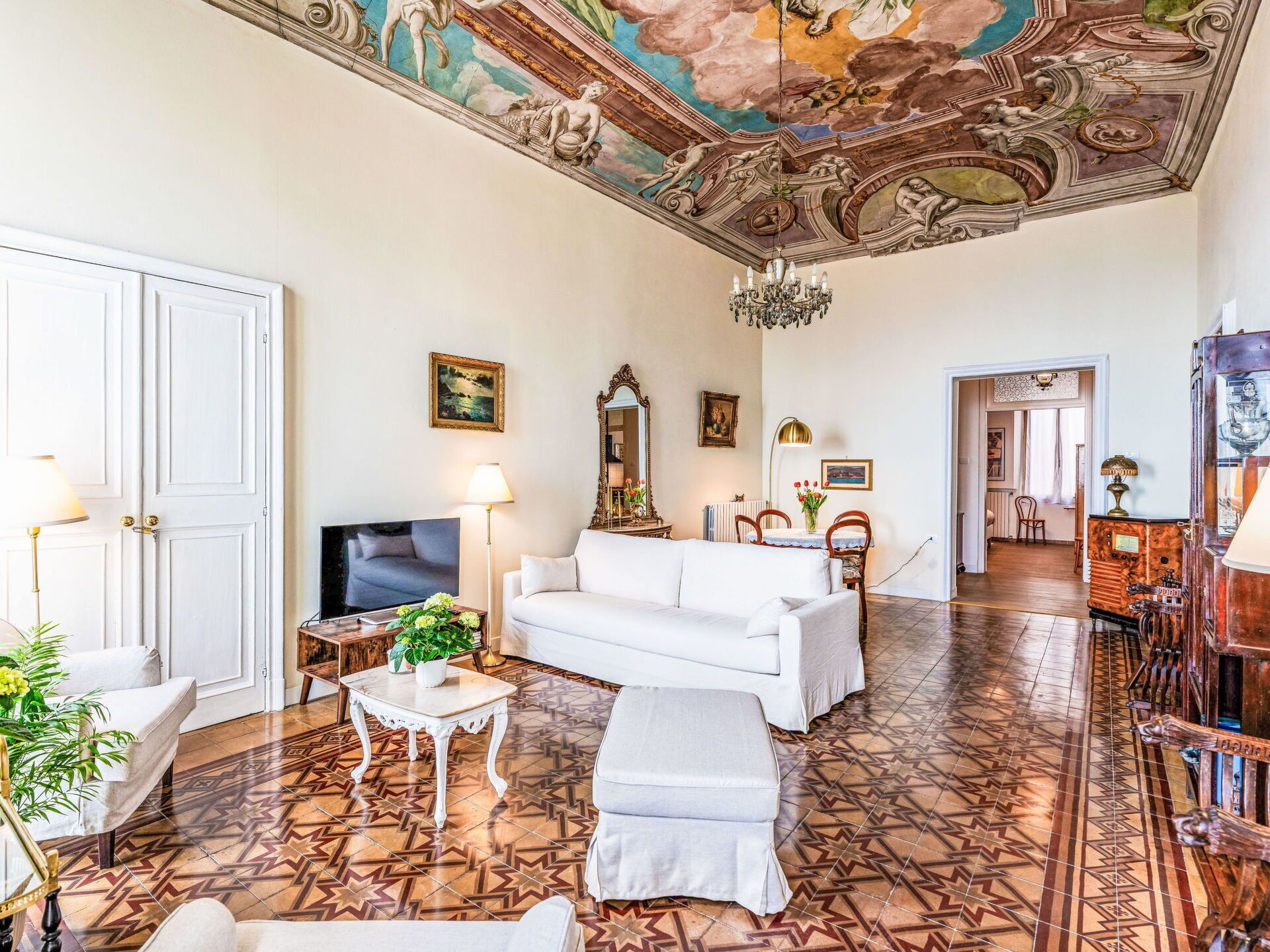 A spacious living room with a white sofa, a television, a chandelier, and a patterned floor.