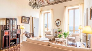 Uma sala de estar ampla com um sofá grande, duas poltronas, uma mesa de centro e um candelabro. Há grandes janelas com cortinas, um mural no teto e um armário com portas de vidro.