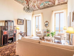 Ein geräumiges Wohnzimmer mit einer großen Couch, zwei Sesseln, einem Couchtisch und einer Chandelieber. Große Fenster mit Vorhängen, ein Deckenfresko und eine Vitrine mit Glastüren sind ebenfalls vorhanden.