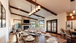 A spacious dining area with a long wooden table set for a meal, a fireplace, and large windows offering a view. - Mayflower Vista - Deer Valley East Basecamp for All Seassons w/ Views, Private Hot Tub (Heber City)