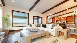 A spacious living area with a large window, a sofa set, a coffee table, and a kitchen in the background. - Mayflower Vista - Deer Valley East Basecamp for All Seassons w/ Views, Private Hot Tub (Heber City)