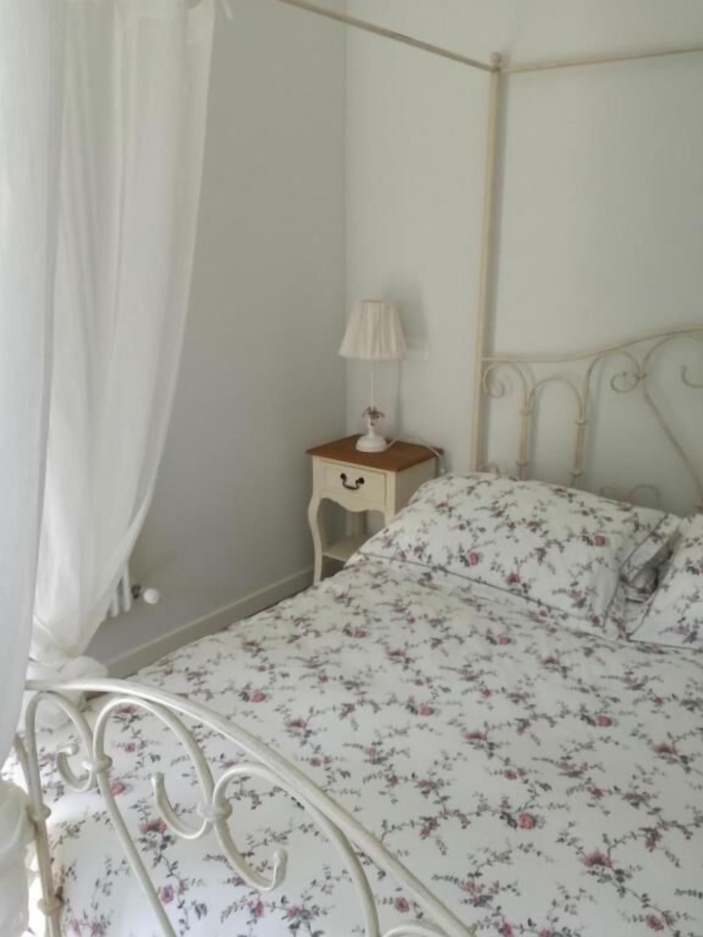 A four-poster bed with a floral bedspread, a bedside table with a lamp, and a white curtain.