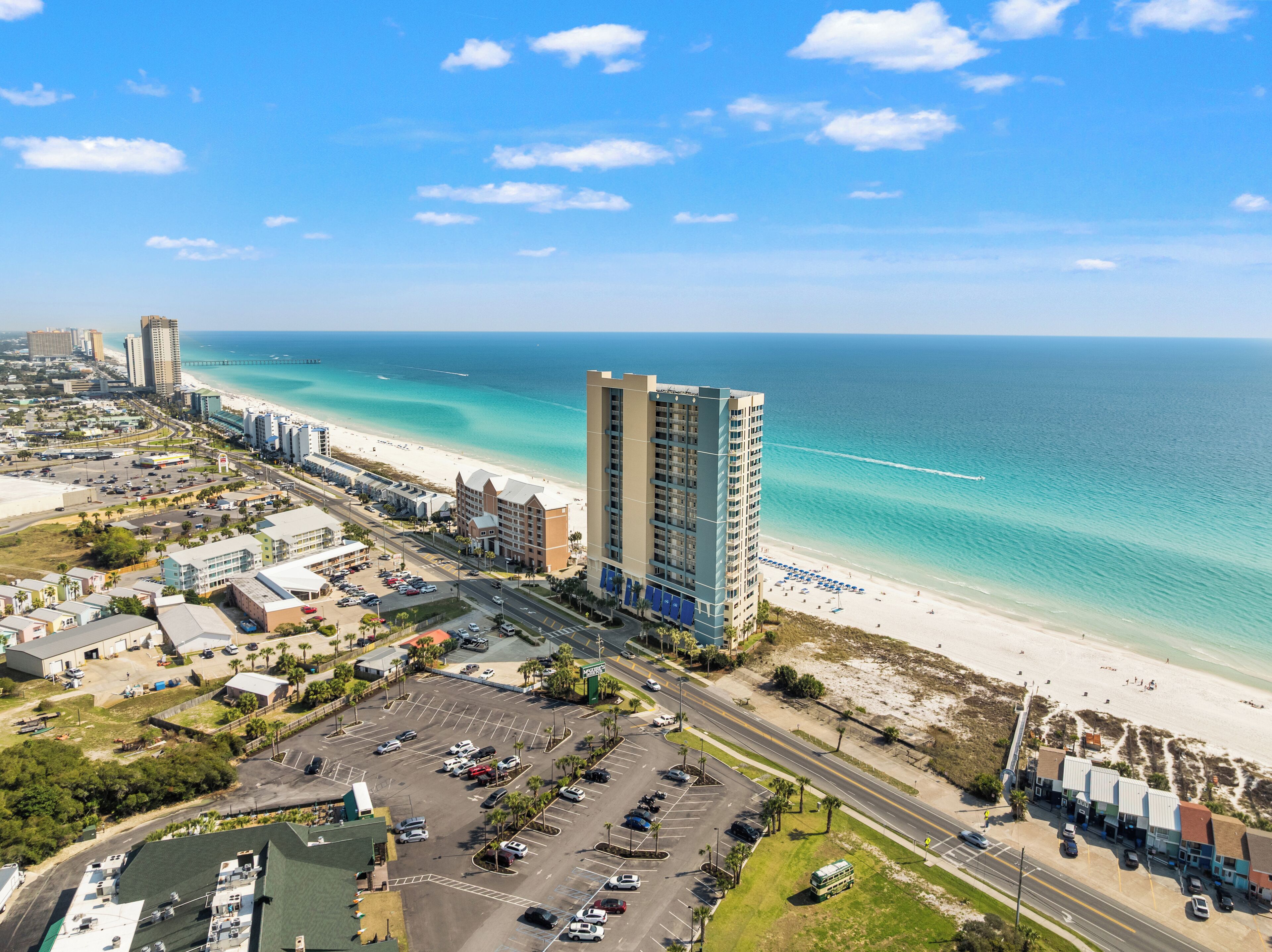 A coastal cityscape with a prominent high-rise hotel, a beach, and a parking area.