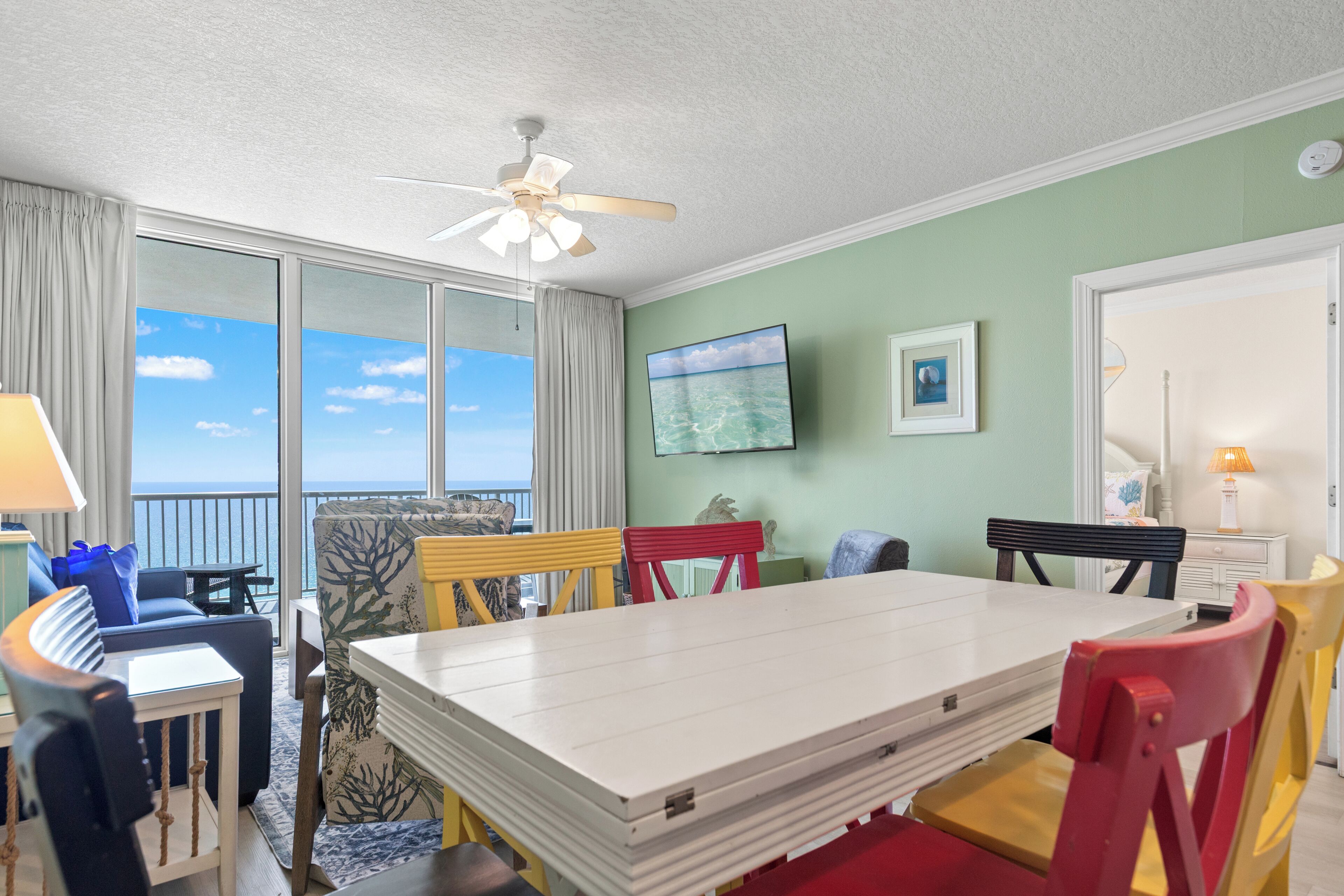 A room with a large window, a ceiling fan, a flat-screen TV, and a dining table with chairs.