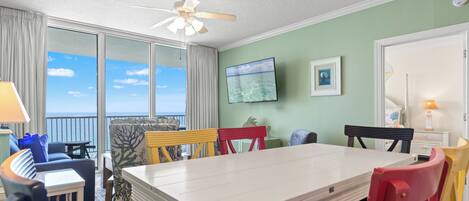 A room with a large window, a ceiling fan, a flat-screen TV, and a dining table with chairs.