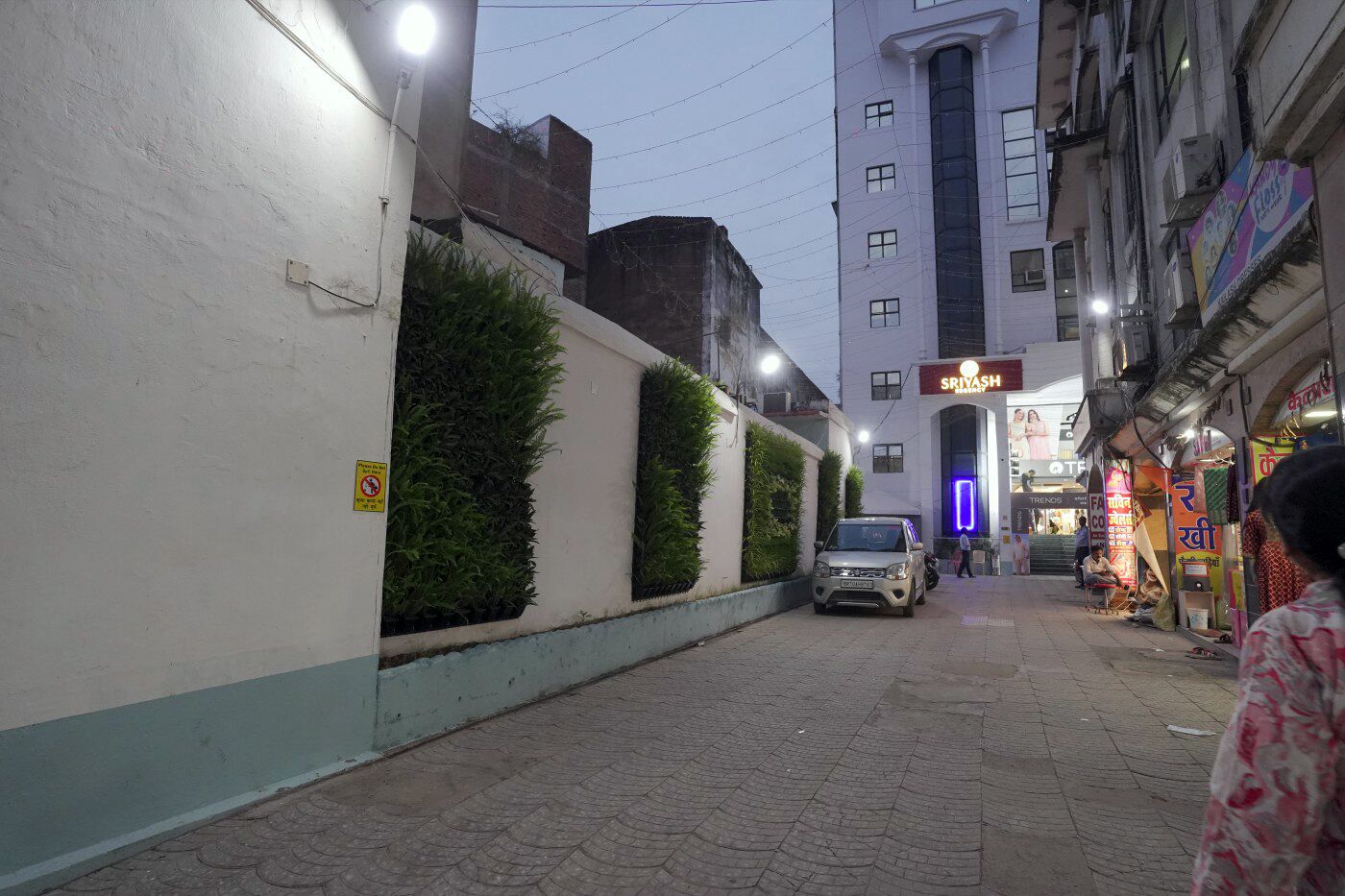 A narrow street with a parked car, a building with a sign reading "SRIYASH," and a shop with a neon sign.