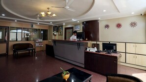 A hotel reception area with a receptionist, a reception desk, a bench, a small table with a plant, and a ceiling fan. - WORMWOOD SYNERGY SRIYASH REGENCY (Bhagalpur)