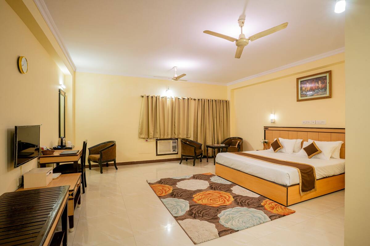 A hotel room with a bed, desk, chairs, a television, and a ceiling fan.