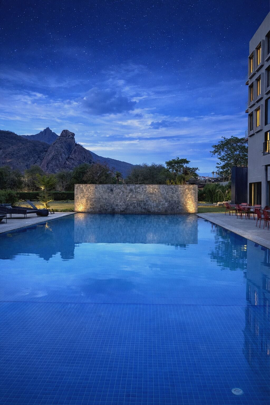 A swimming pool with a stone wall, a mountain in the background, and a building with windows.