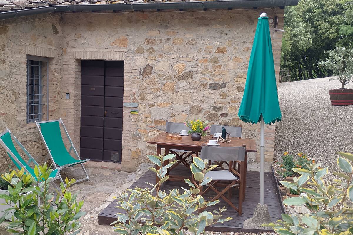 Een gebouw van steen met een houten tafel en stoelen, een groene parasol en potplanten.