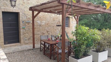 Un edificio de piedra con una pérgola de madera, una mesa con sillas y plantas en macetas.