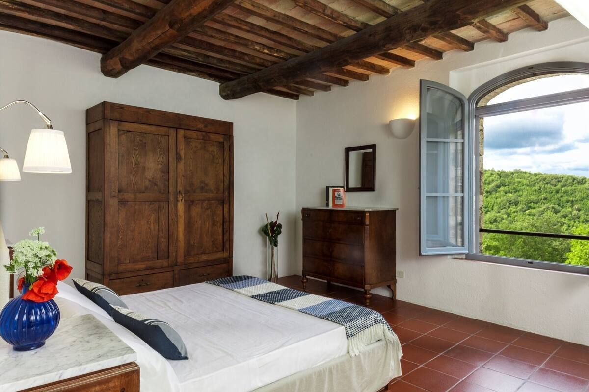 Een slaapkamer met een bed, een houten garderobe, nachtkastje en een raam met uitzicht op groen.