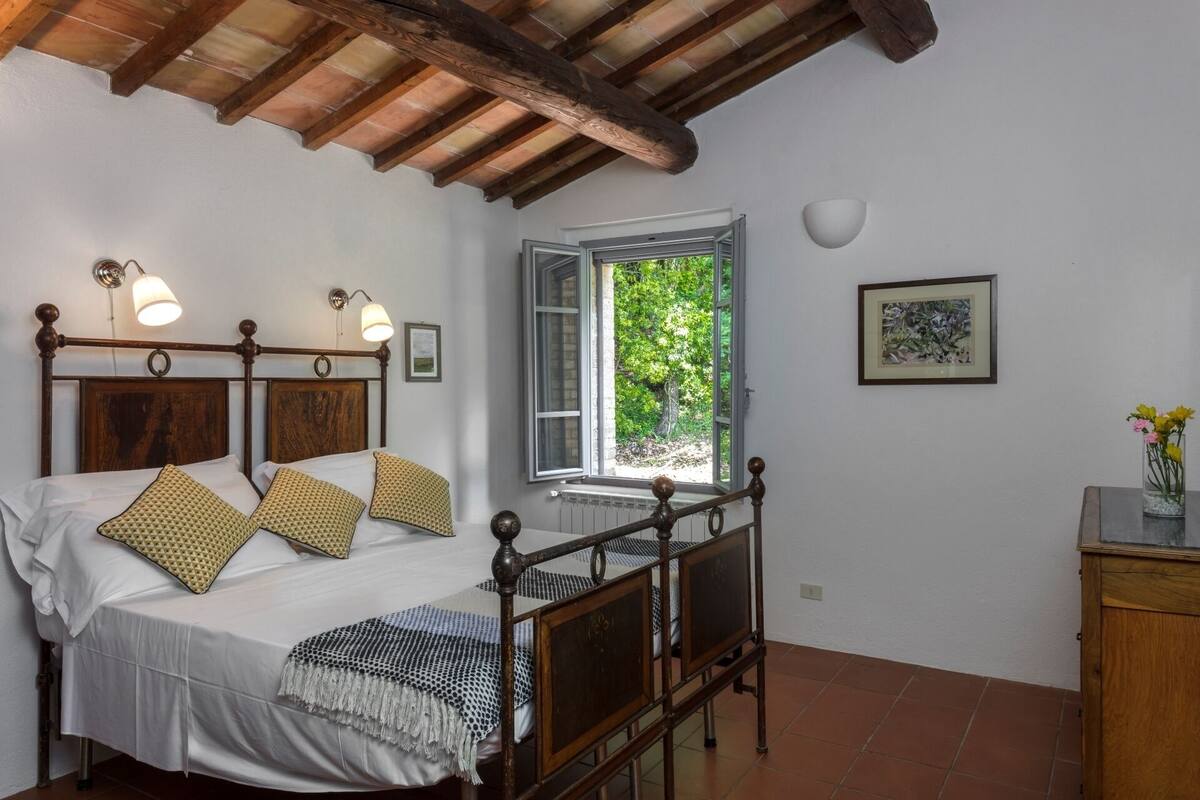 Een slaapkamer met een houten bed, een raam met uitzicht op groen, een ingelijst schilderij aan de muur en een kastje met bloemen.