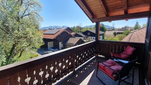 A balcony with a view of houses and mountains, a wooden railing, and a seating area with cushions. - Sunny top-floor apartment with panoramic balcony & mountain views (Greiling)