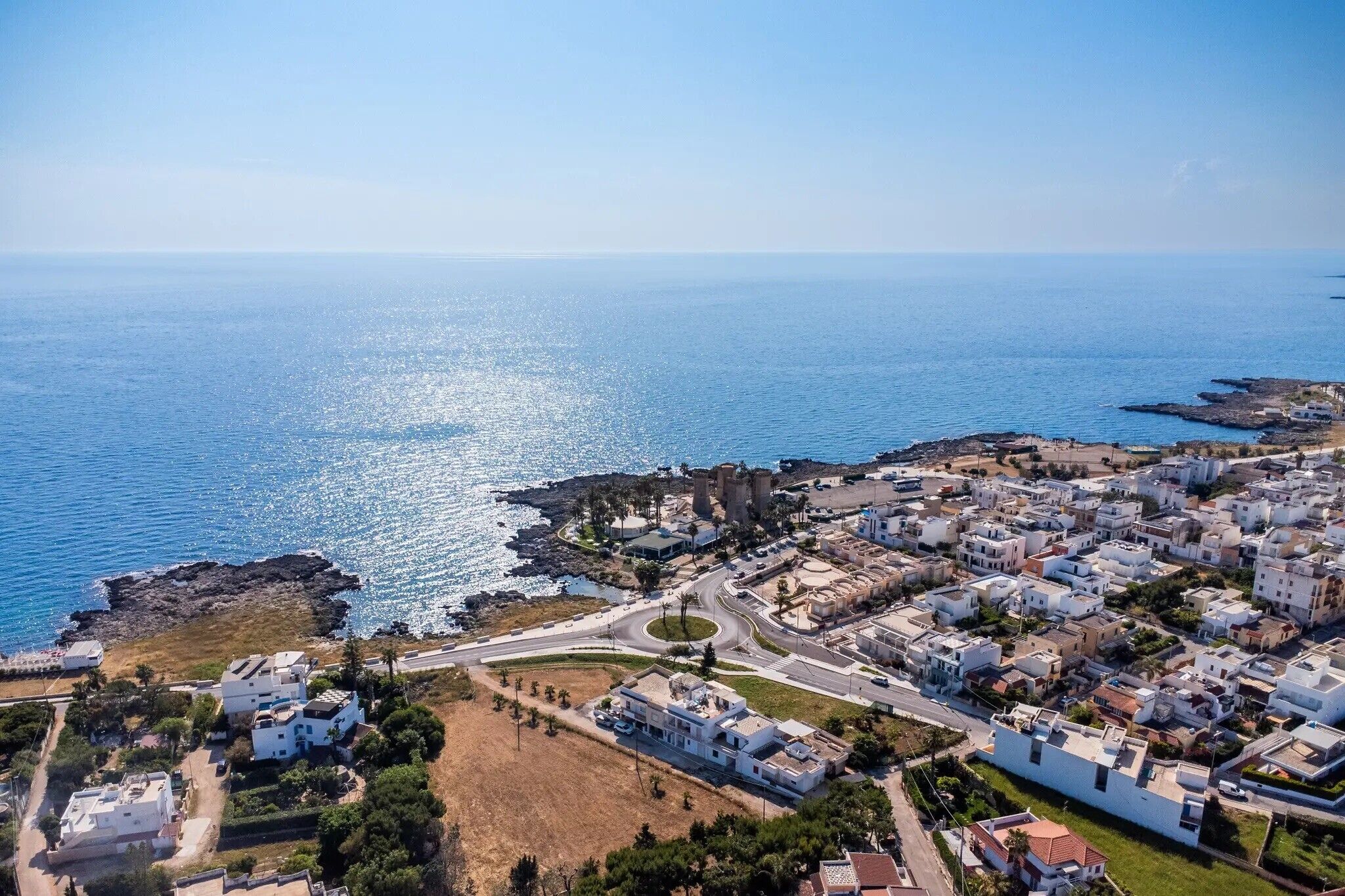 An aerial view of a coastal town with a mix of residential buildings, a prominent castle-like structure, and a clear blue sea.