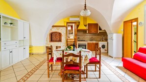 Un coin repas avec une table dressée pour quatre personnes, entouré de chaises en bois avec des coussins rouges, un lustre et une cuisine en arrière-plan.