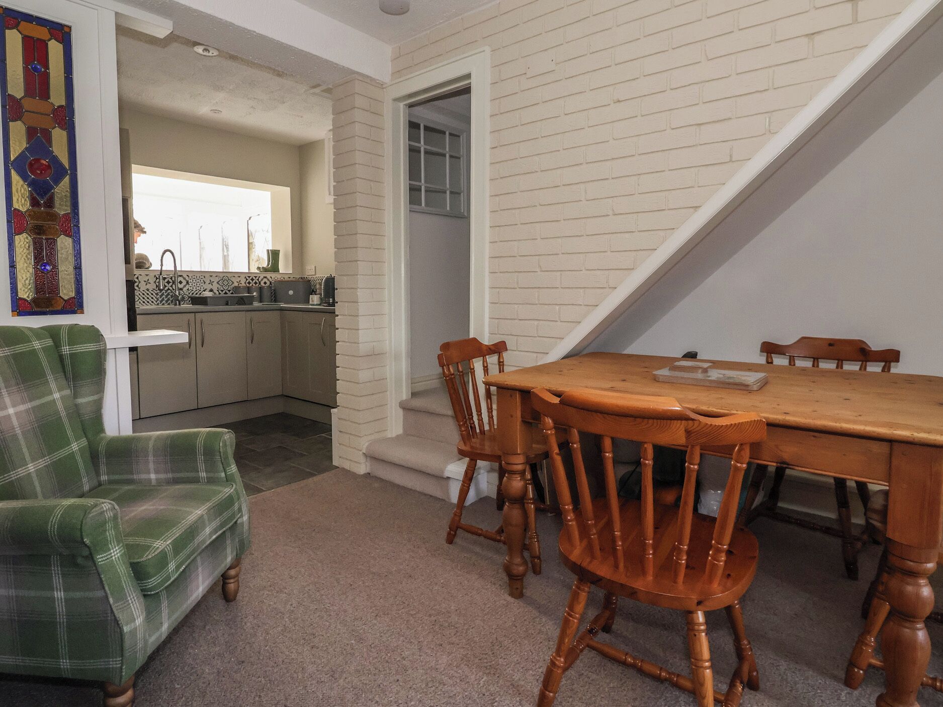 A dining area with a wooden table and chairs, a plaid armchair, and a kitchen in the background.