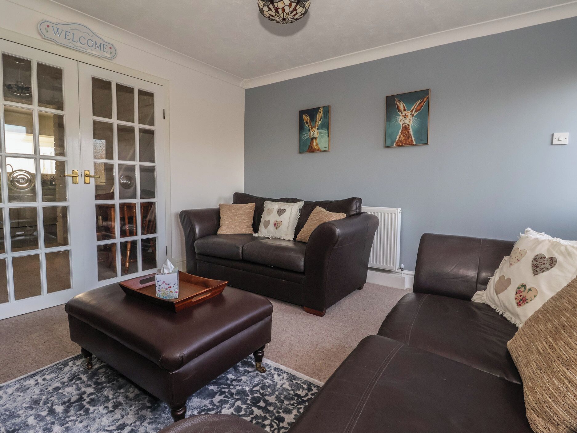 A living room with a dark brown leather sofa, a matching ottoman, and a coffee table. There are two paintings on the wall, a radiator, and a 'Welcome' sign above a door with glass panels.