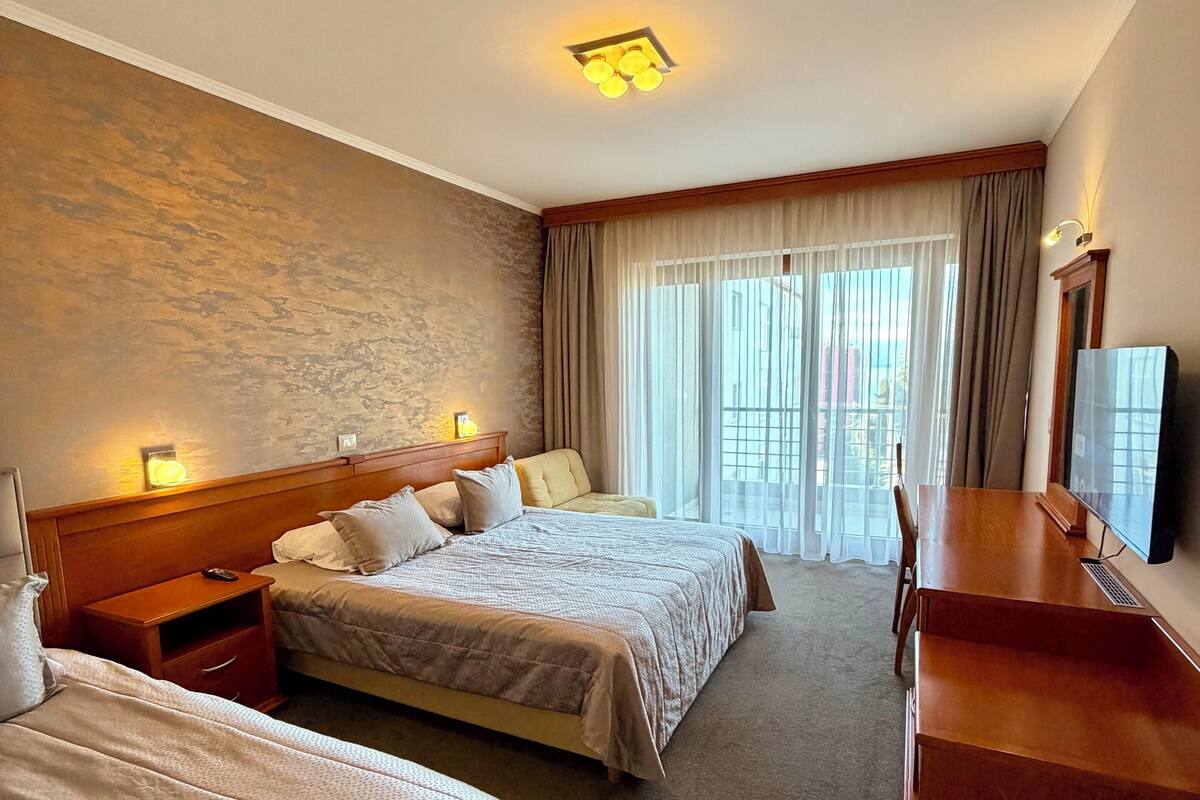 Ein Hotelzimmer mit zwei Betten, einem Schreibtisch, einem Fernseher und einem Balkon mit Vorhängen.
