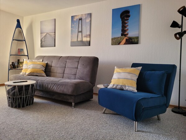 Ein Wohnzimmer mit einem grauen Sofa, einem blauen Sessel und einem kleinen runden Tisch. An der Wand hängen drei gerahmte Bilder.