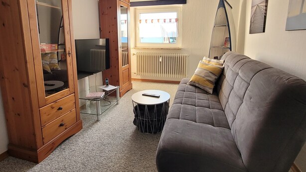 A living room with a sofa, a wooden cabinet, a round coffee table, a TV, and a window with curtains. - Holiday Apartment Zwischenwasser (Jübek)