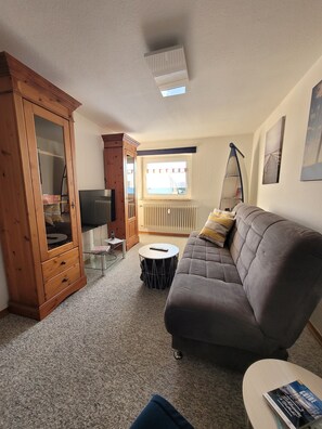 Ein Wohnzimmer mit einer Couch, einem Holzkasten, einem runden Couchtisch, einem Fernseher und einem Fenster mit Vorhängen.
