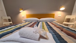 Una cama bien hecha con un edredón colorido, dos almohadas blancas y una toalla suave y blanca con un adorno. Hay dos sillas blancas con un diseño peculiar a los lados de la cama.
