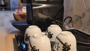 Una encimera de cocina con una cafetera negra, una bandeja con cuatro tazas blancas y una cesta con una tarjeta.