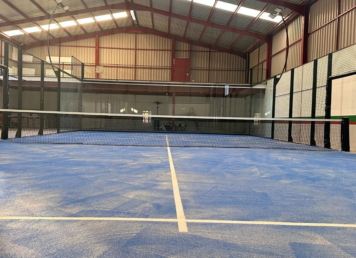 Ein Hallen-Tennisplatz mit Netz, umgeben von Metallwänden und einer hohen Decke.