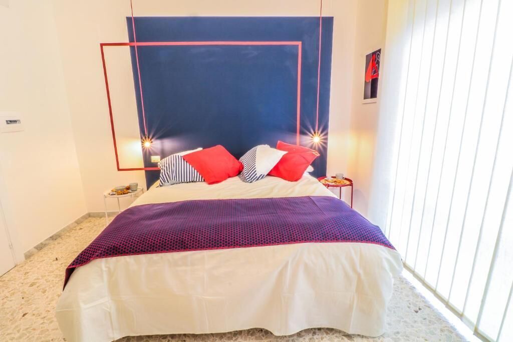 Una cama bien hecha con una colcha blanca y morada, almohadas rojas y blancas, y un cabecero azul.