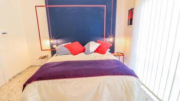 Una cama bien hecha con una colcha blanca y morada, almohadas rojas y blancas, y un cabecero azul.