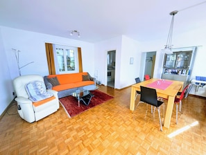 A modern living room with a sofa, a dining table, and a kitchen area visible through an open door.