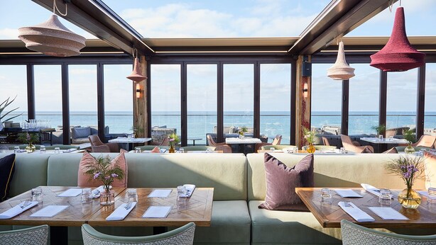 En restaurang med utsikt över havet, inredd med träbord, bekväma sittplatser och hängande lampor.