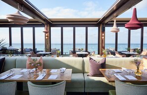 En restaurant med havudsigt, med træborde, polstrede siddepladser og hængende pendellamper.