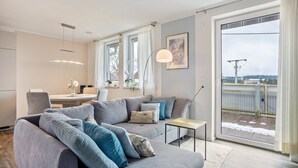 Ein modernes Wohnzimmer mit einem grauen Sofa, einem kleinen Tisch und einem großen Fenster mit Blick auf einen Balkon.